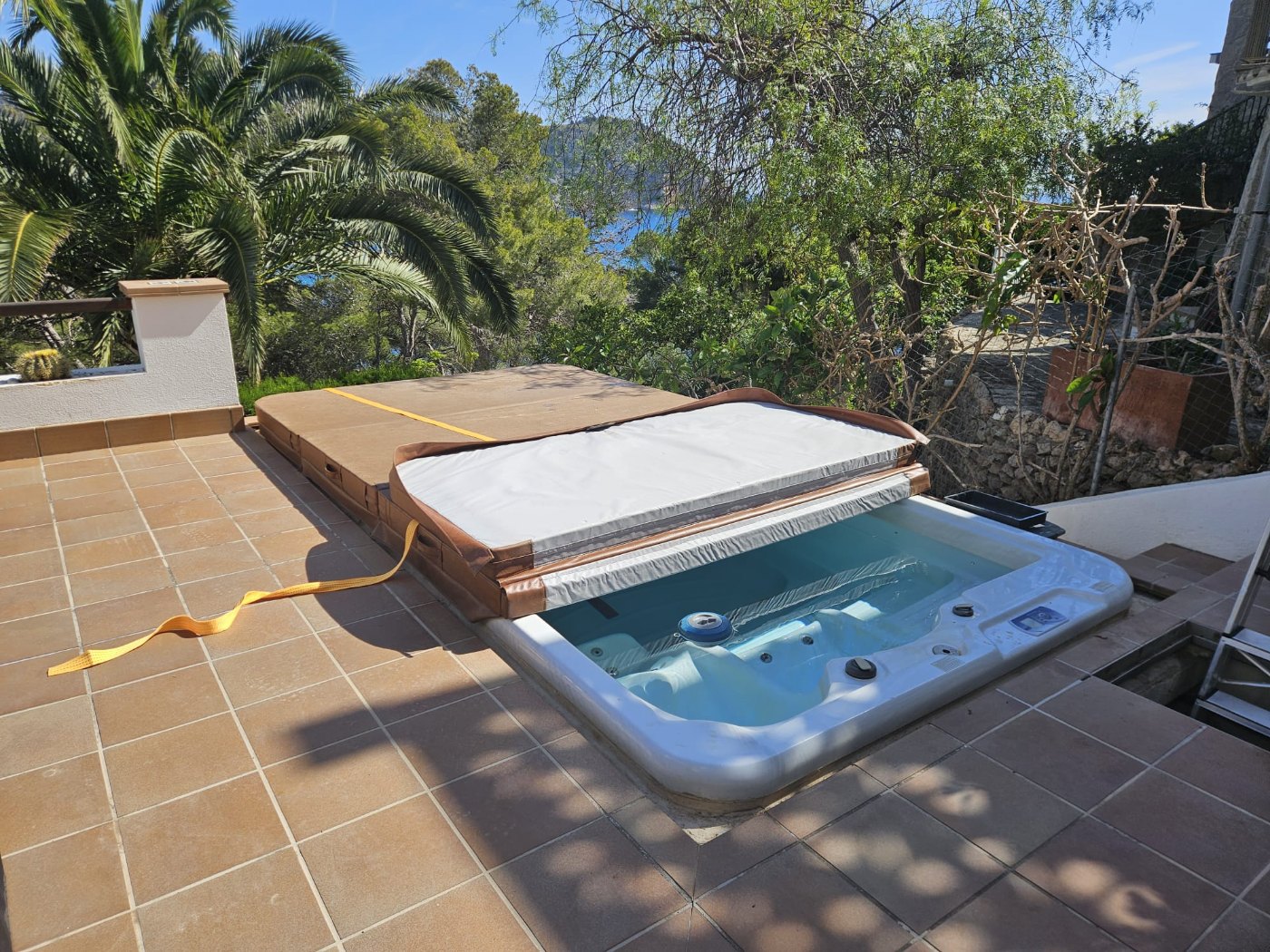 The spa filled and back in service on its tiled terrace in Costa Canyamel, the cover folded back and the bay visible beyond the garden